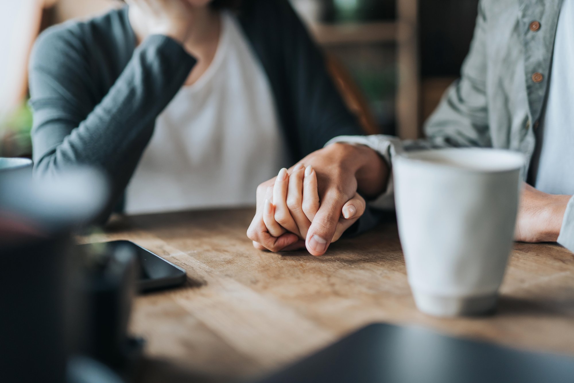Close up of young Asian couple on a date in cafe, holding hands on coffee table. Two cups of coffee and smartphone on wooden table.  