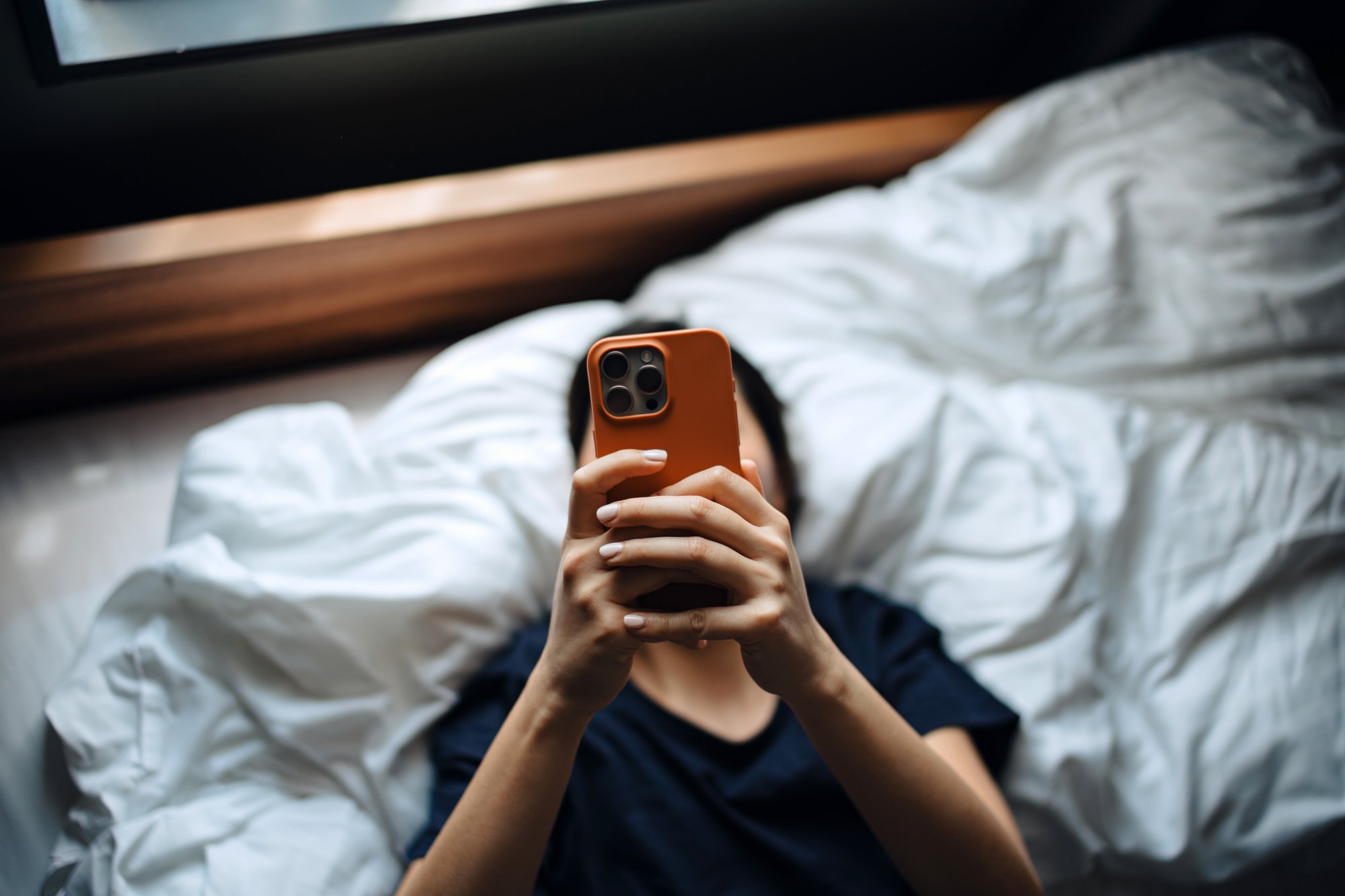 High angle shot of young Asian woman texting on her smartphone while lying on bed relaxing